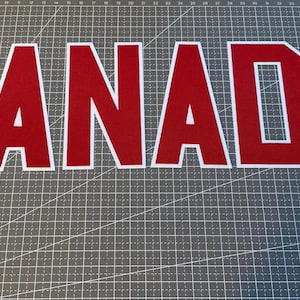 May include: The word CANADA is displayed in large, bold, red block letters with a thin white outline. Each letter features a sharp, angular varsity-style font. The letters are arranged horizontally and are positioned against a dark grey grid mat.