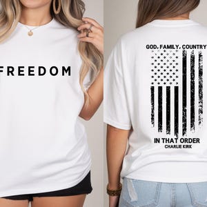 May include: White t-shirt with the word "FREEDOM" in large black letters on the front. The back of the shirt features a distressed American flag design with the text "GOD, FAMILY, COUNTRY IN THAT ORDER CHARLIE KIRK."