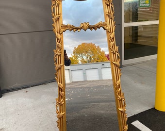 May include: A tall, ornate gold-leaf mirror with a decorative, carved frame. The mirror has a rectangular shape with a slightly arched top and a decorative, carved bottom.