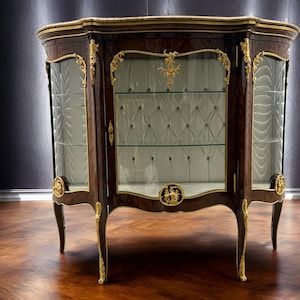 May include: A dark wood display cabinet with gold accents. The cabinet has glass doors, a marble top, and a tufted interior. Ornate gold detailing adorns the corners and legs. The cabinet is a classic piece of furniture.