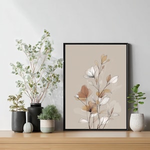 Könnte beinhalten: Ein gerahmter Kunstdruck mit floralem Motiv, der einen beigefarbenen Hintergrund mit filigranen Linienzeichnungen von Blumen in Weiß, Braun und Gold zeigt. Das Kunstwerk befindet sich in einem schwarzen Rahmen und wird auf einer Holzoberfläche mit dekorativen Pflanzen präsentiert.