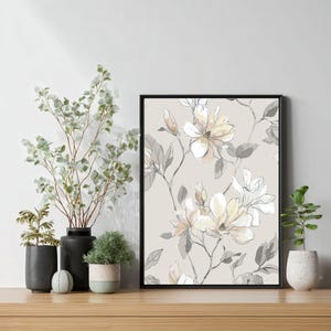 Könnte beinhalten: Ein gerahmter Blumendruck mit einem sich wiederholenden Muster aus Magnolienblüten in sanften Creme-, Weiß- und Grautönen. Das Kunstwerk ist vor einem beigen Hintergrund platziert und mit einem schwarzen Rahmen versehen. Der Druck wird auf einer Holzoberfläche ausgestellt.