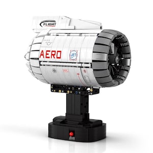 Puede incluir: Un modelo de motor a reacción blanco y gris con la palabra "AERO" en letras rojas. El modelo tiene una base negra y un botón rojo. La palabra "FLIGHT" está impresa en la parte superior del modelo.