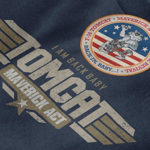 May include: Navy blue garment featuring distressed text and graphics. The design includes the words "TOMCAT" and "MAVERICK ACT" with a star. A circular patch shows a cartoon cat with a gun, the American flag, and the text "F-14 TOMCAT MAVERICK ACT HERITAGE REVIVAL".