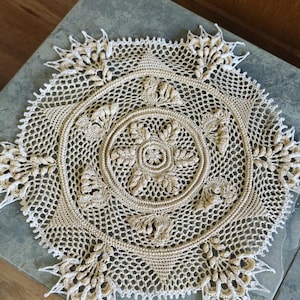 May include: A beige and white crocheted doily with a detailed floral pattern in the center and around the edges. The doily is circular and rests on a gray surface.