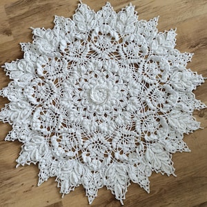 May include: A white, crocheted doily with a detailed floral and leaf pattern. The circular design features a central flower motif and a scalloped edge. The handmade textile is displayed on a wooden surface.