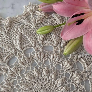 Puede incluir: Un tapete de ganchillo beige hecho a mano con un patrón floral detallado. Flores de lirio rosa y capullos verdes están dispuestas a un lado. El intrincado diseño del tapete y las flores crean una estética decorativa delicada.