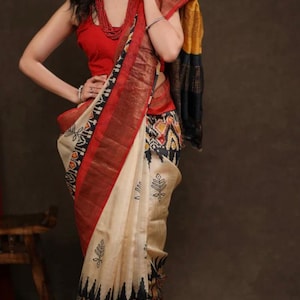 May include: A cream-colored saree featuring a bold red border and black ikat-style geometric patterns. The garment includes a sleeveless red blouse and a draped pallu with black and mustard yellow sections, accented by small black leaf motifs.