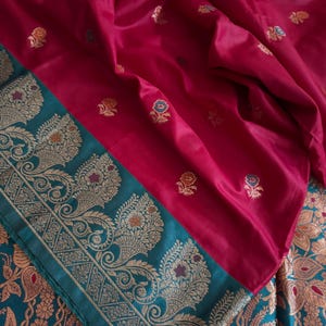 May include: A magenta fabric featuring small, scattered floral motifs in gold and blue. The fabric has a wide, dark teal border decorated with intricate, repeating gold paisley and floral patterns, creating a distinct contrast in color and design.