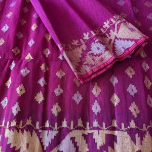 Pode incluir: Um tecido magenta vibrante com um padrão de diamante repetido. O tecido apresenta uma borda decorativa com desenhos geométricos dourados e prateados. A seção dobrada revela uma borda detalhada, mostrando o intrincado artesanato do tecido.