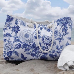 May include: A white canvas tote bag with a blue and white floral pattern. The bag has rope handles and is sitting on a sandy beach.