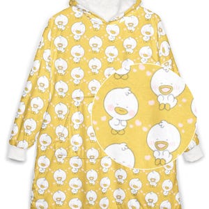 K&ouml;nnte beinhalten: Gelbe Kapuzen-Decke mit einem sich wiederholenden Muster aus wei&szlig;en Cartoon-Enten mit rosa Herzen. Die Decke hat ein wei&szlig;es Fleecefutter in der Kapuze und an den Manschetten. Eine Nahaufnahme zeigt das Enten-Design.