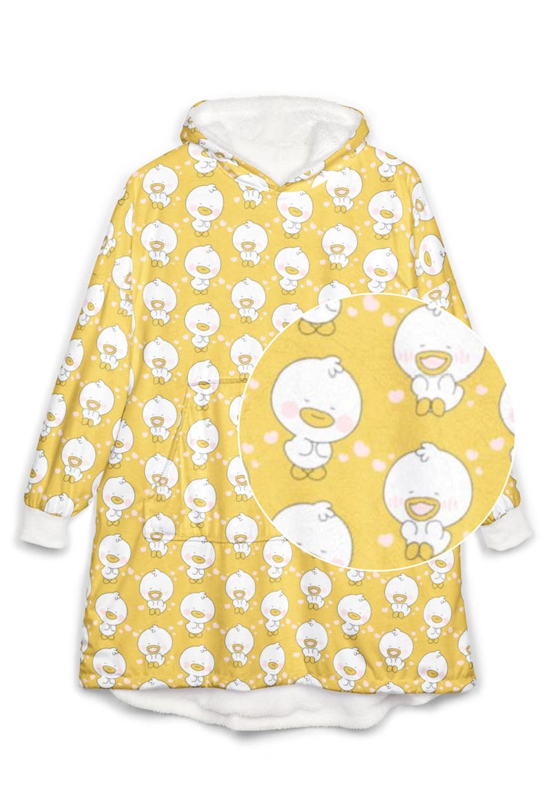 K&ouml;nnte beinhalten: Gelbe Kapuzen-Decke mit einem sich wiederholenden Muster aus wei&szlig;en Cartoon-Enten mit rosa Herzen. Die Decke hat ein wei&szlig;es Fleecefutter in der Kapuze und an den Manschetten. Eine Nahaufnahme zeigt das Enten-Design.