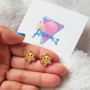 May include: A pair of gold wooden earrings featuring the symbol of the "Sol Inca" or "Inca Sun", a traditional symbol of the Inca Empire. The earrings are on a white card with the word "Radicals" printed on it.
