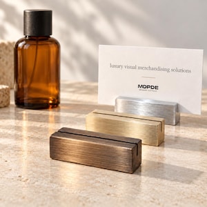 Könnte beinhalten: Drei rechteckige Kartenhalter aus Metall in gebürstetem Bronze-, Gold- und Silber-Finish, jeweils mit einem mittigen Schlitz. Eine weiße Visitenkarte mit dem Text "luxury visual merchandising solutions" und "MDPDE" steckt im silbernen Halter.