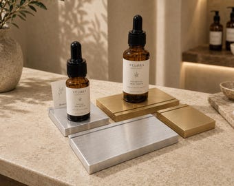 May include: Two amber glass dropper bottles labeled Velora Botanicals, one containing Facial Oil Elixir and the other Botanical Skin Serum, displayed on brushed silver and gold metal rectangular stands with integrated card slots.
