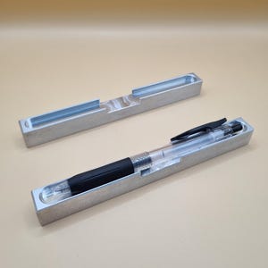 Könnte beinhalten: Ein klarer Stift mit schwarzem Griff liegt in einem Stifthalter aus gebürstetem Metall. Ein weiterer leerer Halter ist sichtbar. Der Stifthalter ist rechteckig mit einer eingelassenen Rinne. Der Hintergrund ist einfarbig beige.