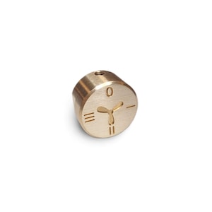 Peut inclure: Objet cylindrique en laiton avec un dessus plat et un corps arrondi. Le dessus présente le chiffre "0", un symbole en forme d'hélice et trois lignes verticales. L'objet a un petit trou sur le dessus et est posé sur un fond blanc.