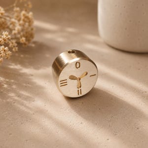 May include: A cylindrical brass-coloured metal object featuring a flat circular face engraved with a central three-petaled floral motif, surrounded by numerical markings including a zero at the top, two vertical lines at the bottom, and horizontal lines.