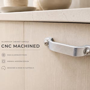 May include: A silver-coloured CNC machined aluminium cabinet handle with a raw finish, attached to a light wood-grain cabinet door with two visible screws. Text on the image reads: ALUMINIUM CABINET HANDLE, CNC MACHINED, RAW ALUMINIUM FINISH, MINIMAL MODERN DESIGN, DESIGNED & MADE IN AUSTRALIA.