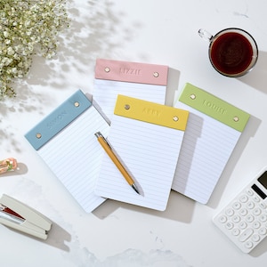 May include: Four personalised lined notebooks with different coloured covers. The covers are light blue, yellow, pink, and green. The notebooks are on a white surface with a calculator, a stapler, a pen, and a cup of coffee.