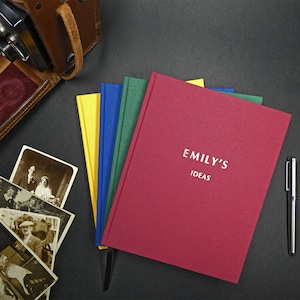 May include: A red hardback journal with the text "Emily's Ideas" in white lettering. The journal is stacked on top of three other journals in yellow, blue, and green.