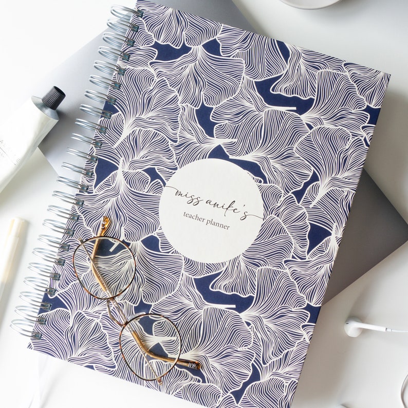 May include: A blue spiral bound teacher planner with a white ginkgo leaf pattern. The cover has a white circle with the text "miss anife's teacher planner".