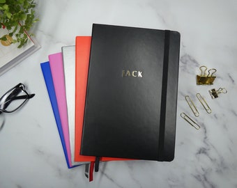 May include: A stack of five notebooks with different colored covers. The top notebook is black with a gold embossed "JACK" on the front. The other notebooks are blue, pink, silver, and red.