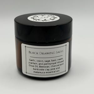 May include: A small, round jar of black drawing salve with a black lid and a white label. The label lists ingredients such as garlic, onion, and essential oils. The jar is on a white background.