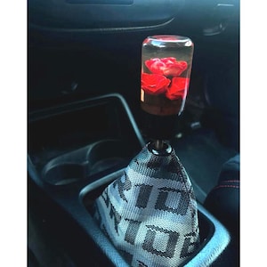 Peut inclure: Un pommeau de levier de vitesses transparent avec des roses rouges à l'intérieur. Le pommeau est monté sur une tige noire, et le soufflet de levier de vitesses est en tissu gris à motifs. L'intérieur de la voiture est visible en arrière-plan.