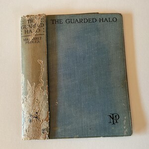 May include: A vintage hardback book titled "The Guarded Halo" by Margaret Pedler. The book cover is blue with a black "NP" logo in the bottom right corner. The spine is worn and faded.