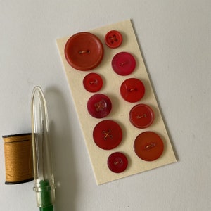 Peut inclure: Assortiment de boutons en plastique rouge et orange de différentes tailles. Les boutons ont une finition brillante et sont disposés sur une carte beige.