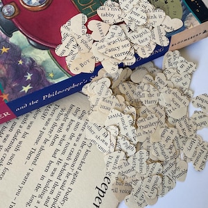 May include: Heart-shaped confetti cut from book pages, scattered near a book with a blue spine. The book's title is "and the Philosopher's". The confetti has text from the book printed on it.