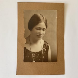 May include: A vintage sepia-toned portrait photograph of a person with a centre-parted hairstyle, wearing a dark sleeveless top with a beaded texture and a long beaded necklace. The photograph is mounted on a rectangular brown cardstock backing.