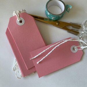 Puede incluir: Una pila de etiquetas de regalo rosas con cuerda blanca y un círculo blanco en el centro. Las etiquetas están sobre una superficie blanca.