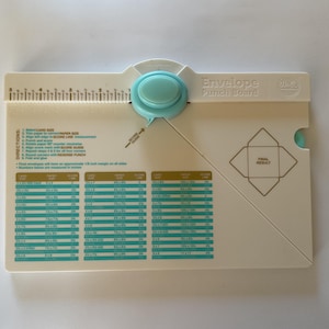 May include: White envelope punch board with a teal knob and ruler markings. The board has printed instructions and a chart for card sizes, paper sizes, and score lines. The text "Envelope Punch Board" is printed on the board.