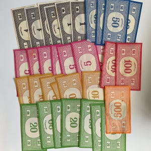 May include: A collection of Monopoly game money in various denominations. The bills are in colors such as gray, pink, orange, green, and blue. Denominations include 1, 5, 10, 20, 50, 100, and 500.