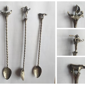 May include: Three silver stirring spoons with decorative handles. The handles feature a small boat, a man with a hat, and a cow. The spoons are all the same size and shape.