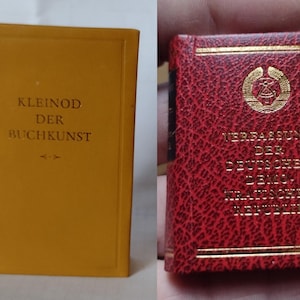 May include: Two small, hardback books. The book on the left has a yellow cover with the text "KLEINOD DER BUCHKUNST" on the front. The book on the right has a red cover with gold lettering that reads "VERFASSUNG DER DEUTSCHEN DEMOKRATISCHEN REPUBLIK".