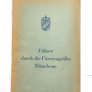 May include: Vintage guide titled "Führer durch die Fürstengrüfte Münchens" in German. The cover is a faded blue-green with a Bavarian coat of arms and crown emblem. The edges show a tan discoloration.