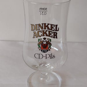 May include: A clear glass beer stein with a gold and brown logo that reads "Dinkel Acker CD-Pils". The logo also features a coat of arms with a wheat sheaf and the letters "CD".