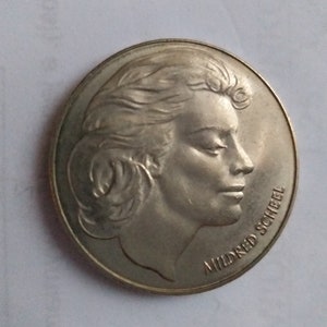 May include: Silver coin featuring a profile portrait of Mildred Scheel. The coin has a smooth, metallic surface.