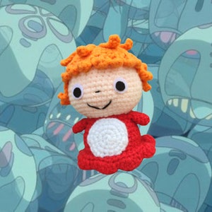 Può includere: Un giocattolo di peluche all'uncinetto con capelli arancioni, un corpo rosso e un cerchio bianco sul petto. Il giocattolo è circondato da uno sfondo blu e bianco.