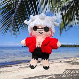 May include: A crocheted plush toy of a character with white hair, wearing a red jacket and black pants. The toy is posed with its arms outstretched and is standing on a beach.