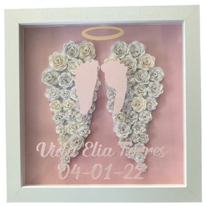May include: A framed artwork featuring angel wings made of white paper flowers, with pink footprints in the center. The background is pink, and the frame is white. The artwork includes the name "Vida Elia Torres" and the date "04-01-22".