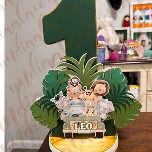 Puede incluir: Un adorno para tarta de cumpleaños con temática de jungla que presenta un gran número uno verde con ribete dorado. El adorno incluye un jeep de safari con el nombre "LEO" y varios personajes de animales, rodeados de hojas verdes.