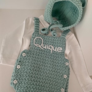 Könnte beinhalten: Ein hellgrünes gehäkeltes Baby-Outfit mit passender Mütze. Das Outfit hat einen weißen gestickten Namen "Quique" auf der Vorderseite und weiße Knöpfe an der Seite. Die Mütze hat Bärenohren.