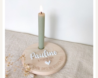 May include: A wooden candle holder with the name "Pauline" and the date "16 Februar 2024" engraved on it. The text "Wie schön, dass du geboren bist!" is also engraved on the holder. A lit green candle is placed in the holder.