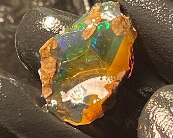 May include: A rough opal gemstone with a vibrant display of colors, including green, blue, and orange. The opal has a textured surface and is held in a black gloved hand. The stone has a natural, raw appearance.