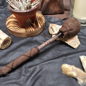 May include: A handcrafted brown leather-wrapped club with a textured head. The handle is wrapped with a darker brown leather. The club rests on a black surface with other rustic items, including a small wooden bowl and animal bones.